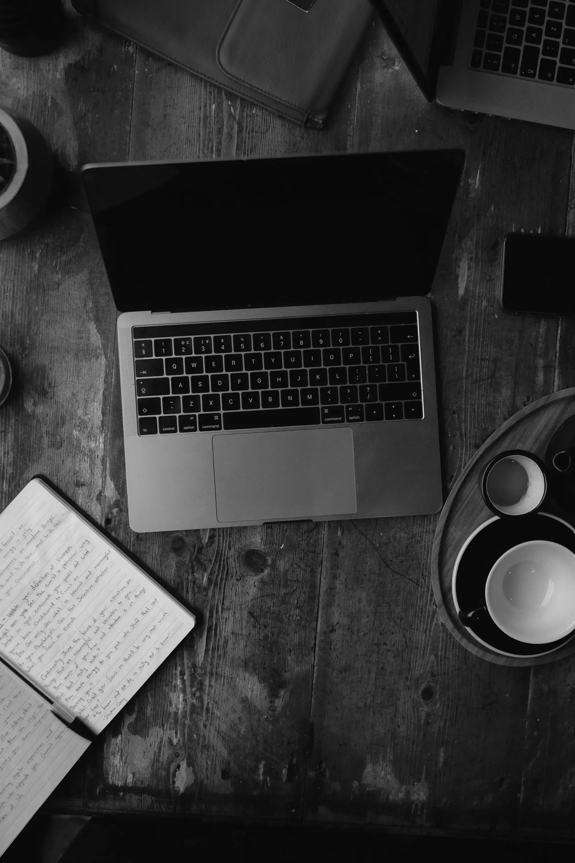 A desk from above with a laptop, notebook and coffee, black and white